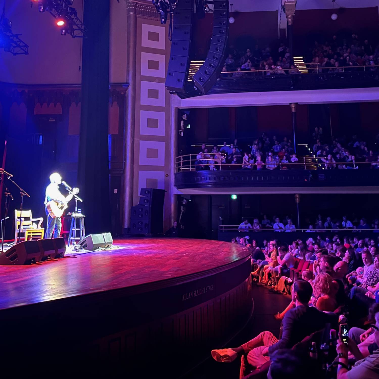 Square picture of an artist with a guitar on a stage facing the audience.